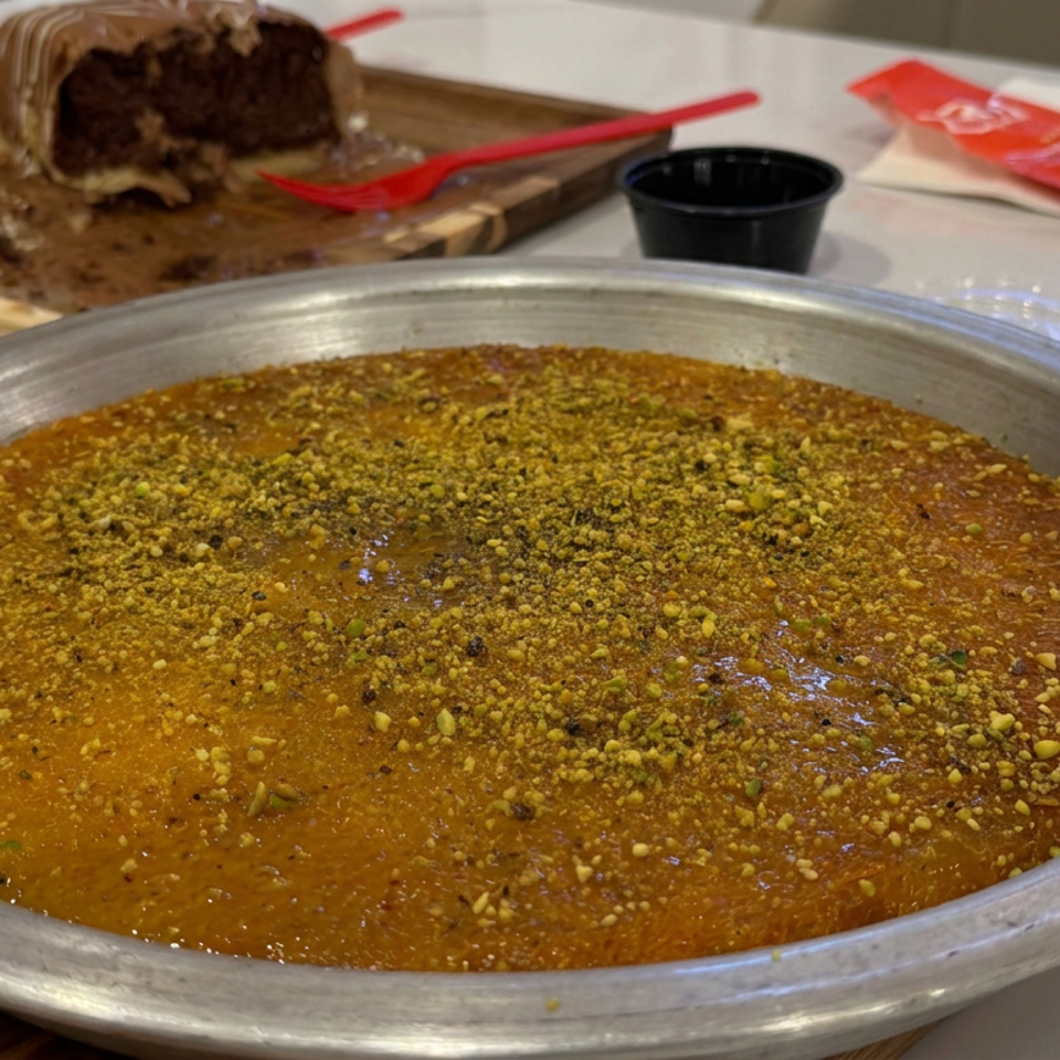 Large round pistachio-topped dessert in a metal serving pan, with sliced chocolate cake, red forks, and dipping sauce behind it.