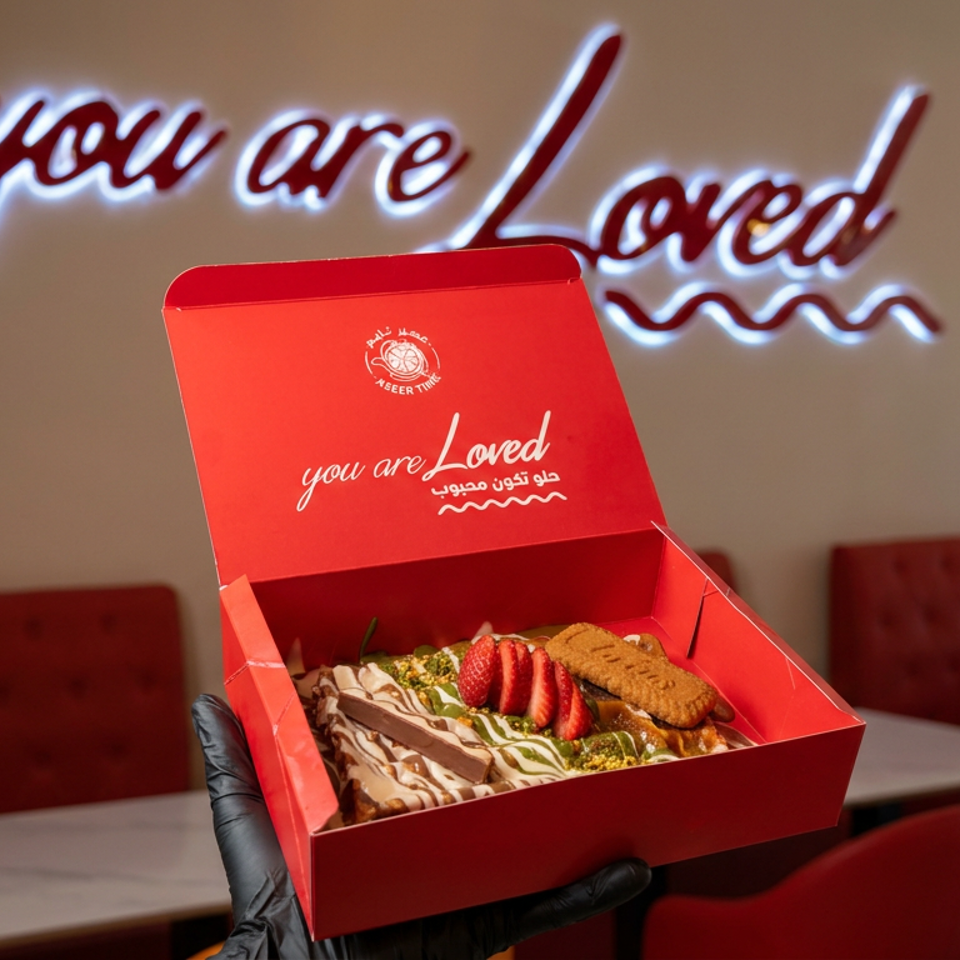Gloved hand presents a red dessert box filled with layered sweets, strawberries, pistachios, and cookies inside a warmly lit restaurant.