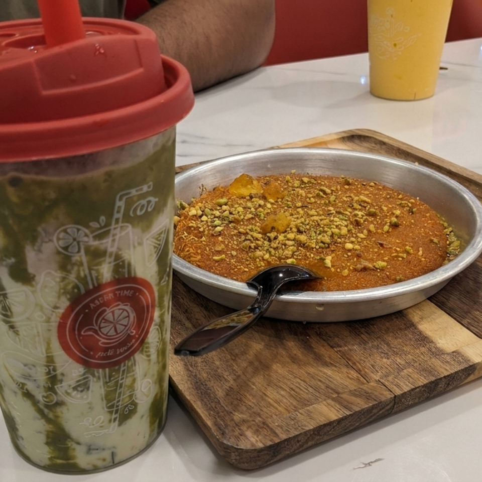 Pistachio-topped dessert in a metal dish with spoon beside a green layered drink on a wooden serving board.