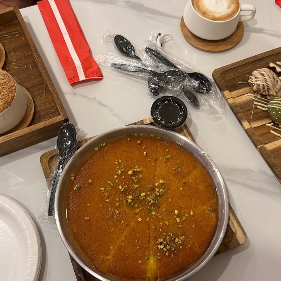 Dessert spread with pistachio-topped cake, decorated pastries, two cappuccinos, and utensils on a marble table.